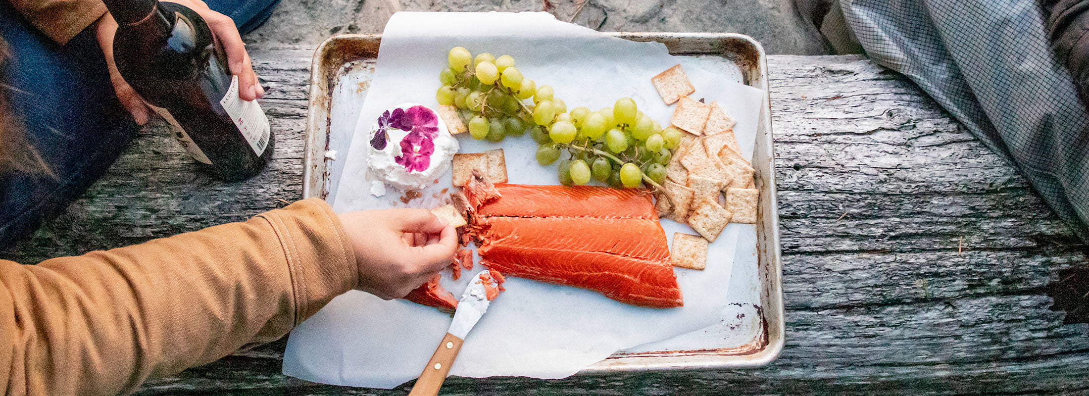 Hands reach out to a beach picnic tray covered with Patagonia Provisions Wild Sockeye Salmon, crackers, fresh grapes and goat cheese